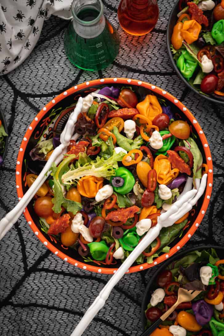 An orange bowl filled with with Halloween salad made with greens, olives, sliced peppers, dyed tortellini, mozzarella cheese skulls and pepperoni cut into bat shapes, with skeleton hands reaching in to serve the salad.