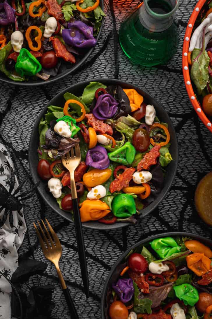 A black plate topped with with Halloween salad made with greens, olives, sliced peppers, dyed tortellini, mozzarella cheese skulls and pepperoni cut into bat shapes, with a fork digging into the salad.