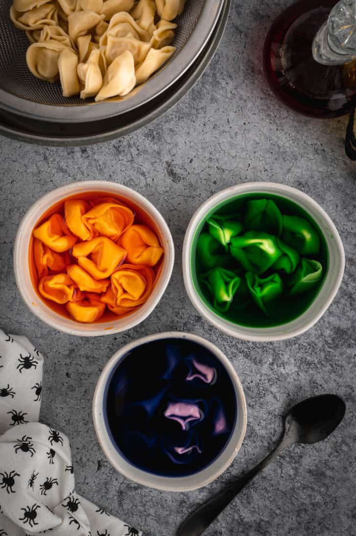 Tortellini soaking in 3 bowls, each filled with dyed water. One bowl has orange water, one has green water, and one has purple water.