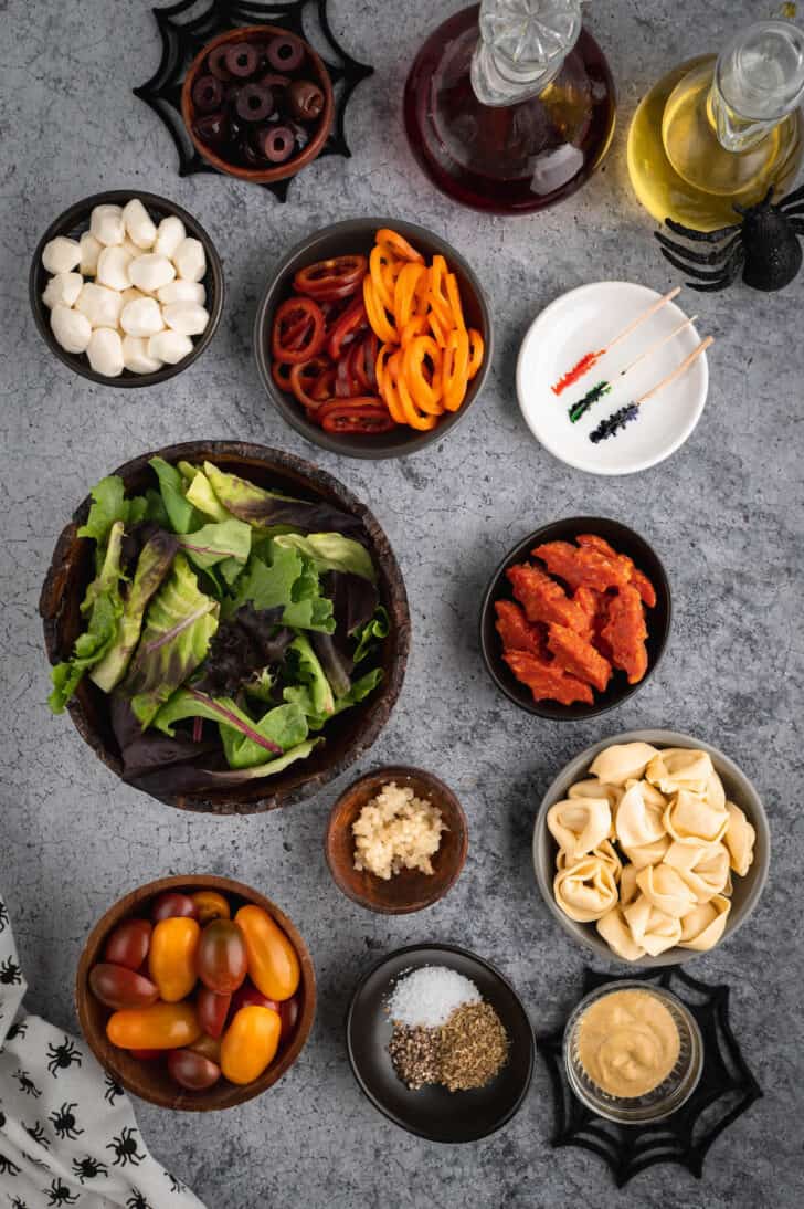 The ingredients for spooky salad on a light gray surface, including mixed greens, sliced peppers, mozzarella cheese balls, pepperoni, tortellini, garlic, tomatoes, mustard and salt and pepper.
