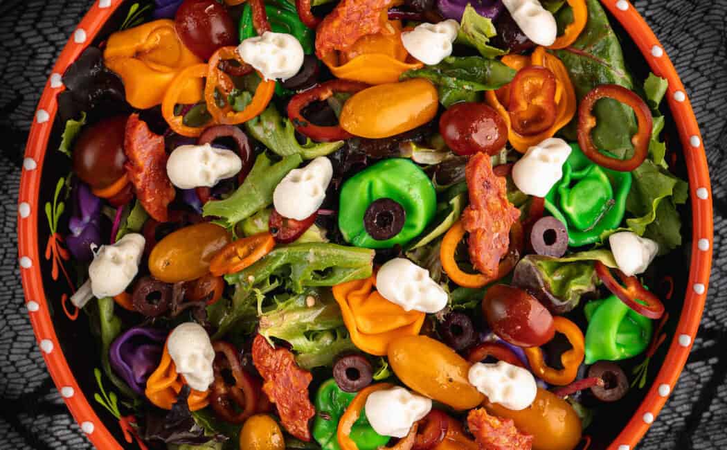 An orange bowl filled with with Halloween salad made with greens, olives, sliced peppers, dyed tortellini, mozzarella cheese skulls and pepperoni cut into bat shapes.