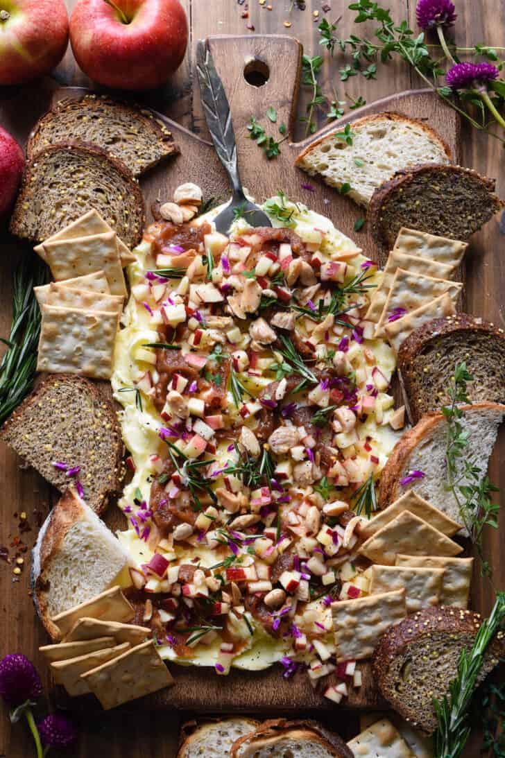 A butter board made with apple butter, fresh apples, nuts and herbs on a wooden cutting board, with bread and crackers for dipping.