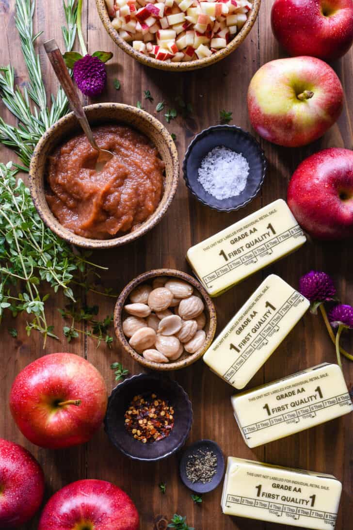The ingredients needed to make an autumn butter board arranged on a wooden surface, including butter, whole and chopped apples, apple butter, red pepper flakes, salt, pepper, marcona almonds, edible flowers, thyme and rosemary.