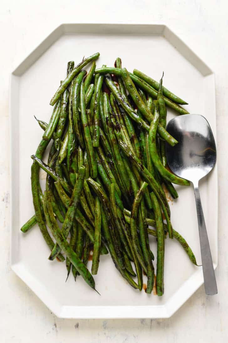 Green beans with balsamic vinegar on a geometric white platter with a silver serving spoon.