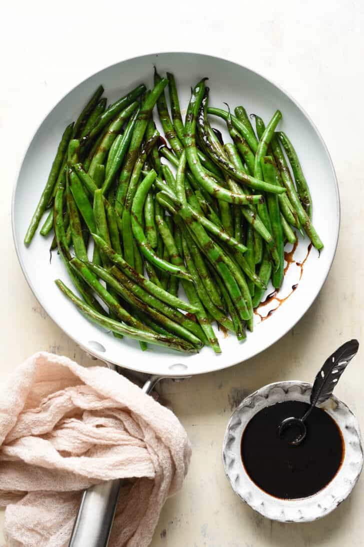 Balsamic green beans in a white skillet, with a pink linen wrapped around the handle, and a small bowl of balsamic glaze in the corner.