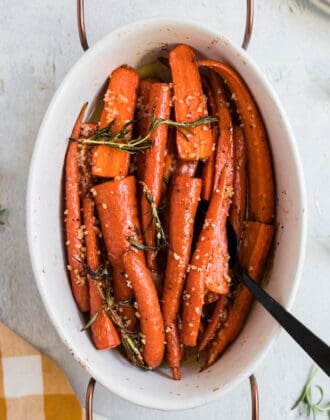 White oval ceramic baking dish with wooden handles filled with garlic honey roasted carrots garnished with rosemary.