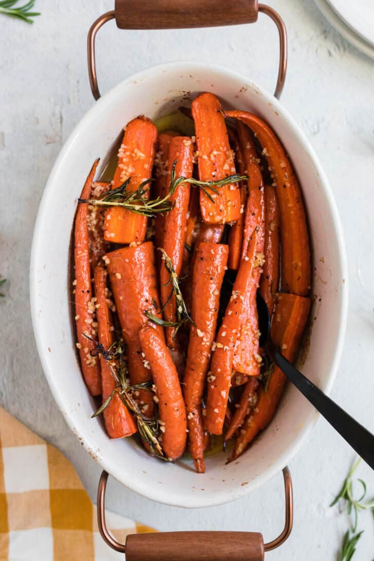 White oval ceramic baking dish with wooden handles filled with honey glazed carrots garnished with rosemary.