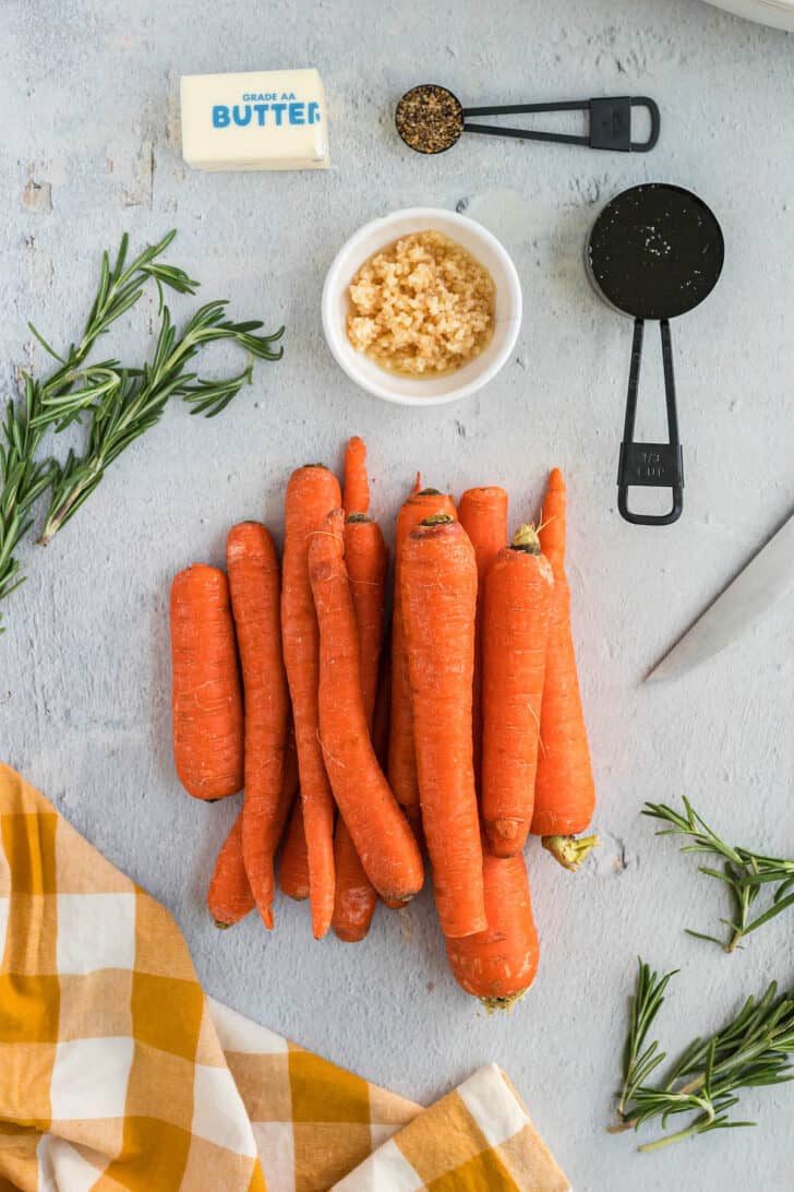 Ingredients for oven glazed carrots arranged on a light surface, including chopped garlic, butter, seasonings and herbs.