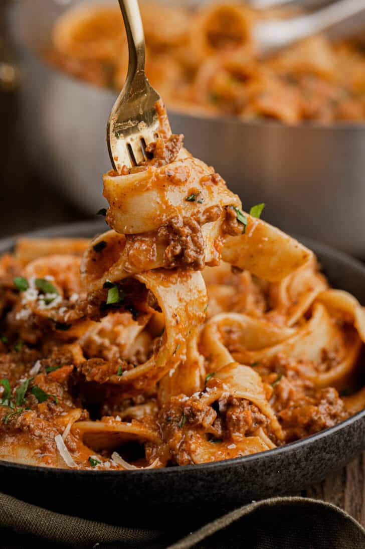 A gold fork twirling pappardelle pasta with lamb bolognese in a black bowl.