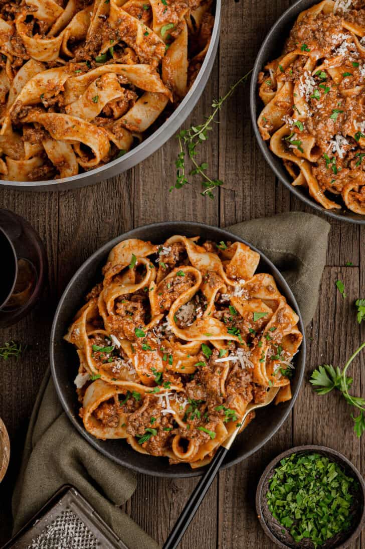 A black bowl filled with ground lamb pasta sauce over pappardelle noodles, garnished with parsley and cheese.