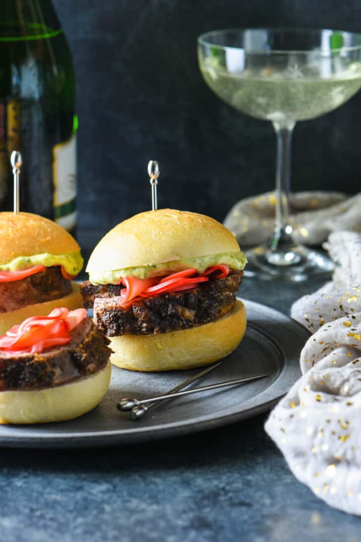 Small sandwiches made with roasted meat on rolls garnished with pickled radishes and pesto mayo, and skewered with decorative silver toothpicks.