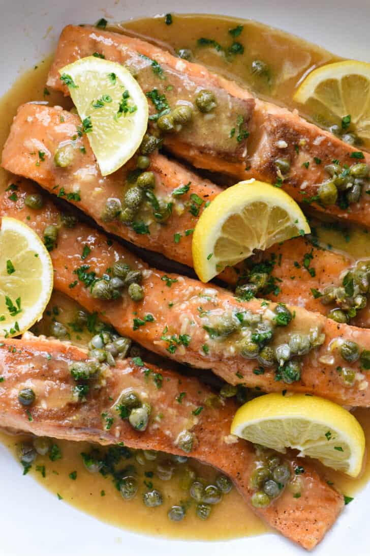 Salmon piccata in a white skillet, garnished with lemon wedges.