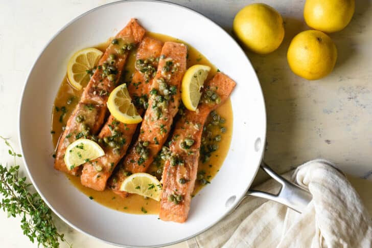 Salmon piccata in a white skillet, garnished with lemon wedges.