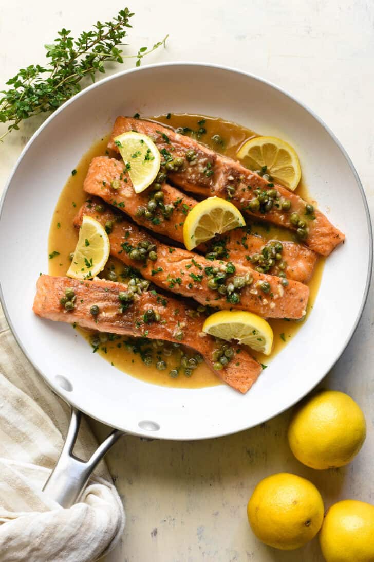 Salmon piccata in a white skillet, garnished with lemon wedges.