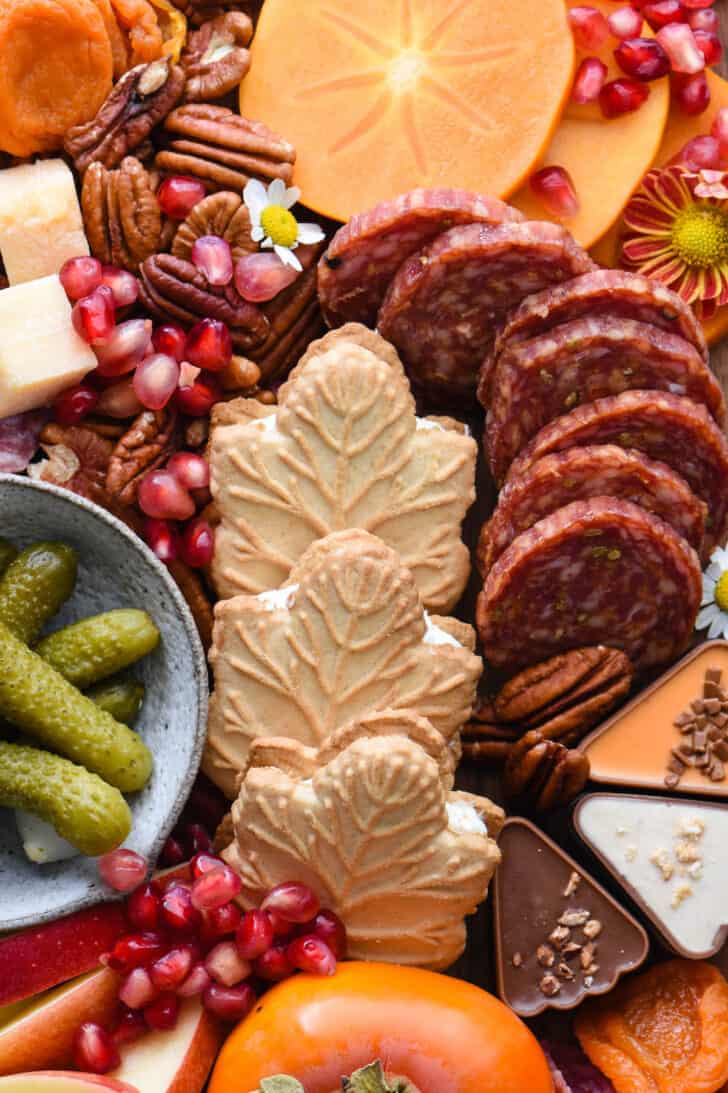 A fall charcuterie board with salami, nuts, pomegranate seeds, pickles and maple leaf cookies.