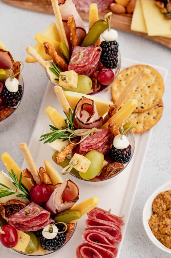 A white platter serving plastic glasses filled with meats, cheeses, fruits, pickles and herbs.