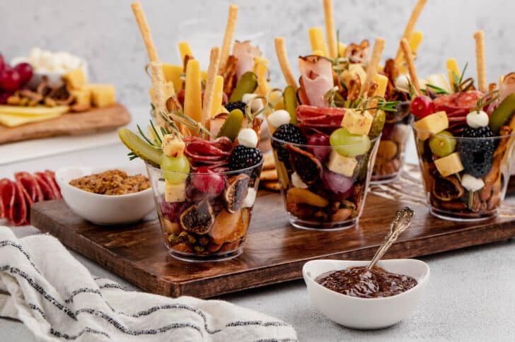 Individual charcuterie cups presented on a wooden cutting board with small bowls of mustard and preserves alongside.