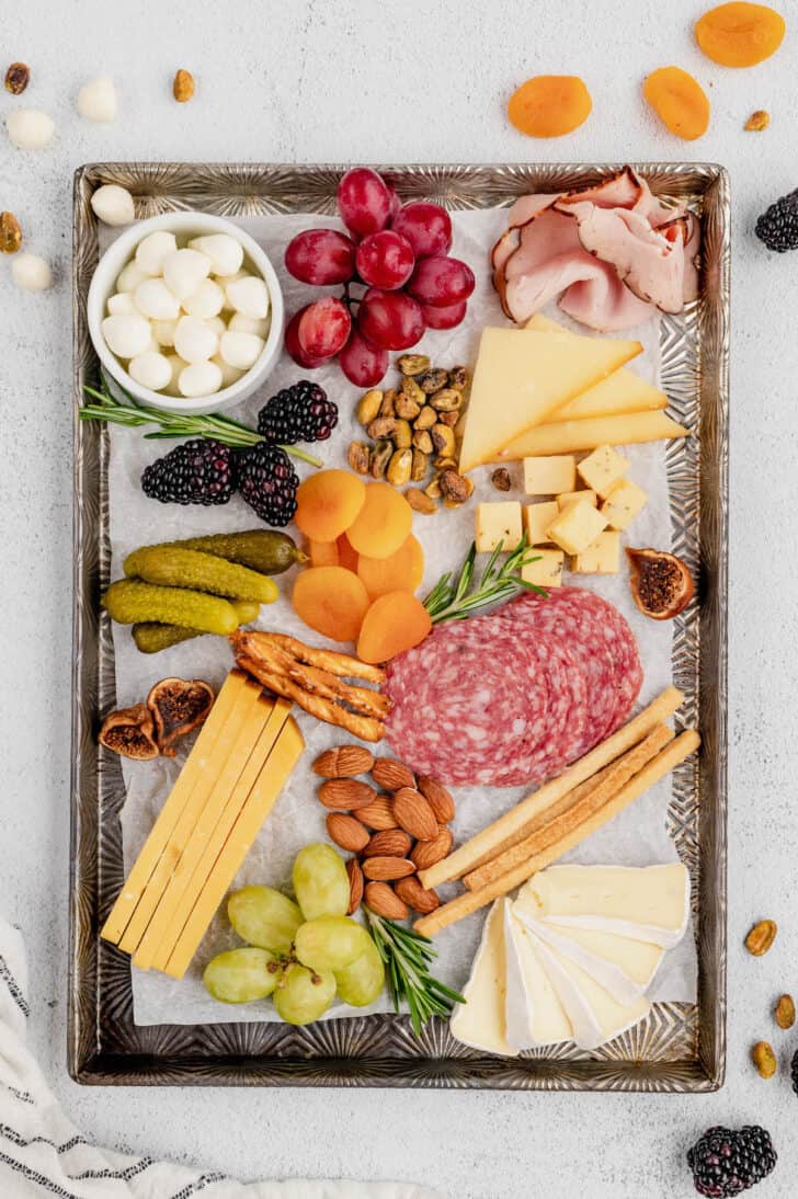 All of the ingredients for individual charcuterie cups on a textured metal tray, including meats, cheeses, fresh and dried fruits, pickles, herbs and nuts.