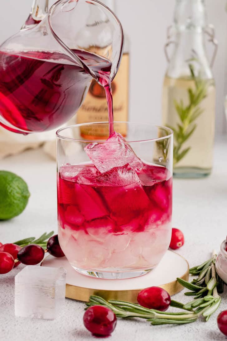 A double old fashioned glass filled with ice, with cranberry juice being poured over the ice.