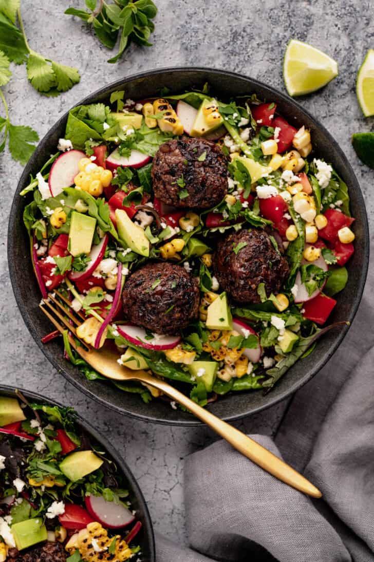 A salad made with greens, chopped tomato, avocado, corn, sliced radishes and cotija cheese topped with meatballs fiber balls.