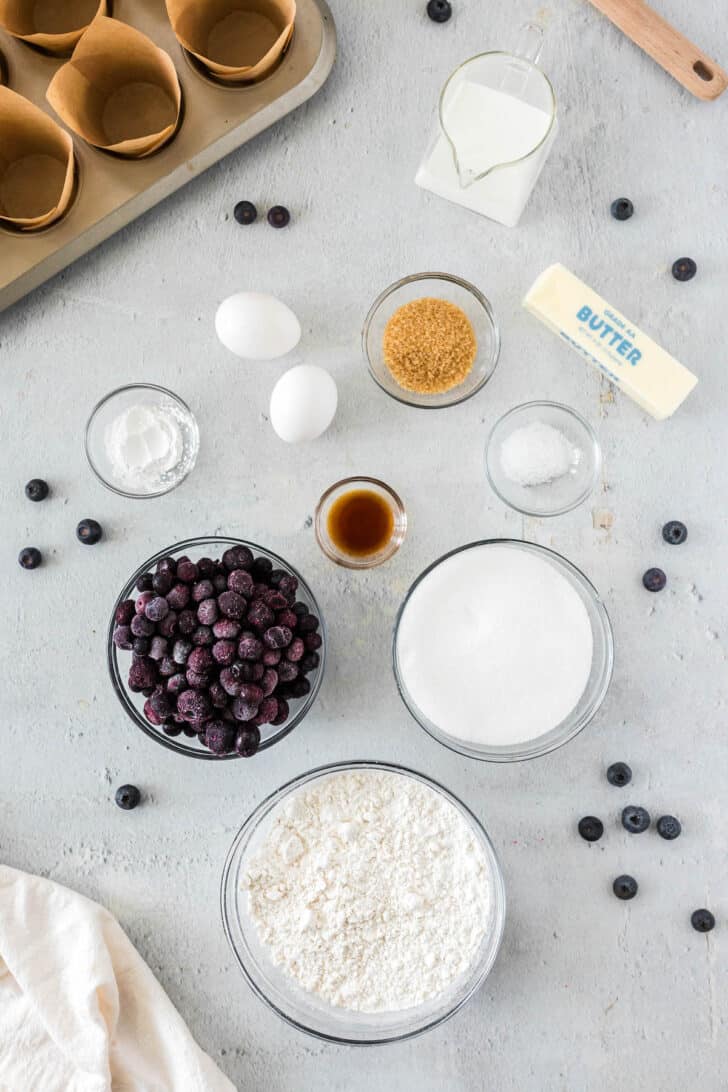 Ingredients on a light surface, including flour, sugar, extracts, blueberries, eggs and butter.
