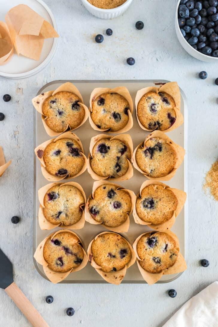Overhead photo of a dozen buttermilk blueberry muffins in brown paper wrappers in a muffin pan.