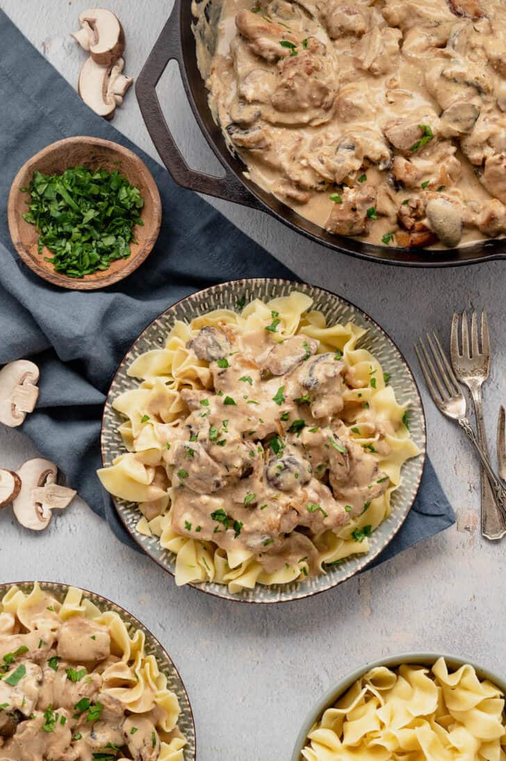 A plate of egg noodles topped with chicken stroganoff, alongside a pan of stroganoff sauce, a bowl of chopped parsley, and extra noodles.