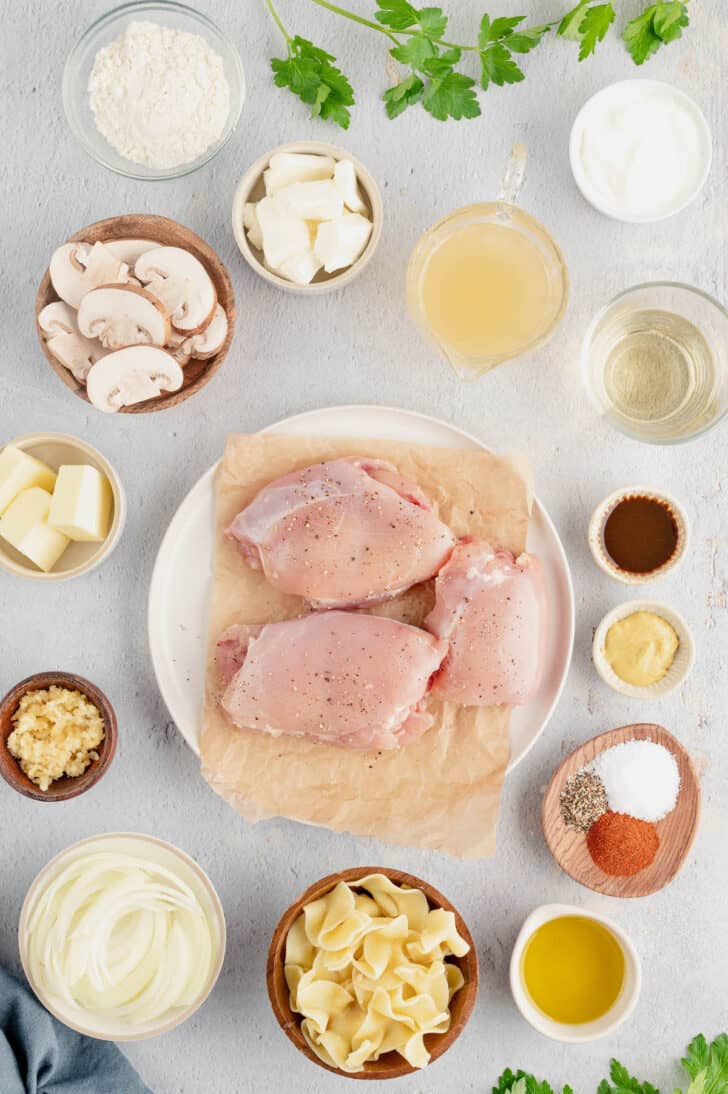 Ingredients laid out on a light surface, including raw poultry, mushrooms, butter, sour cream, cream cheese, condiments and spices.