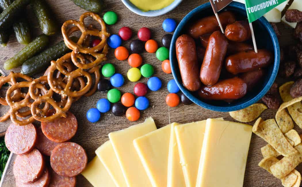 A football charcuterie board made on a wooden cutting board, with cheese, sliced pepperoni, pickles, pretzels, candy, mustard, corn chips and cocktail weenies.