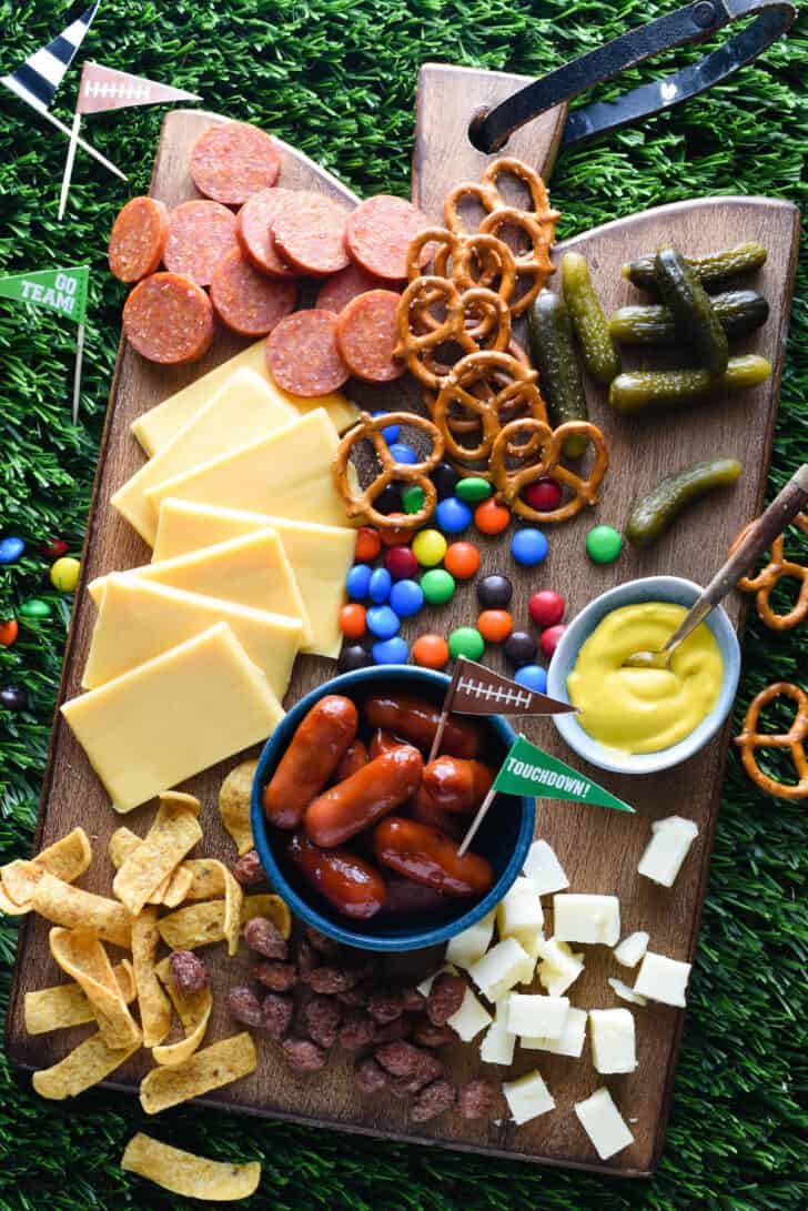 A football charcuterie board made on a wooden cutting board, with cheese, sliced pepperoni, pickles, pretzels, candy, mustard, corn chips and cocktail weenies.