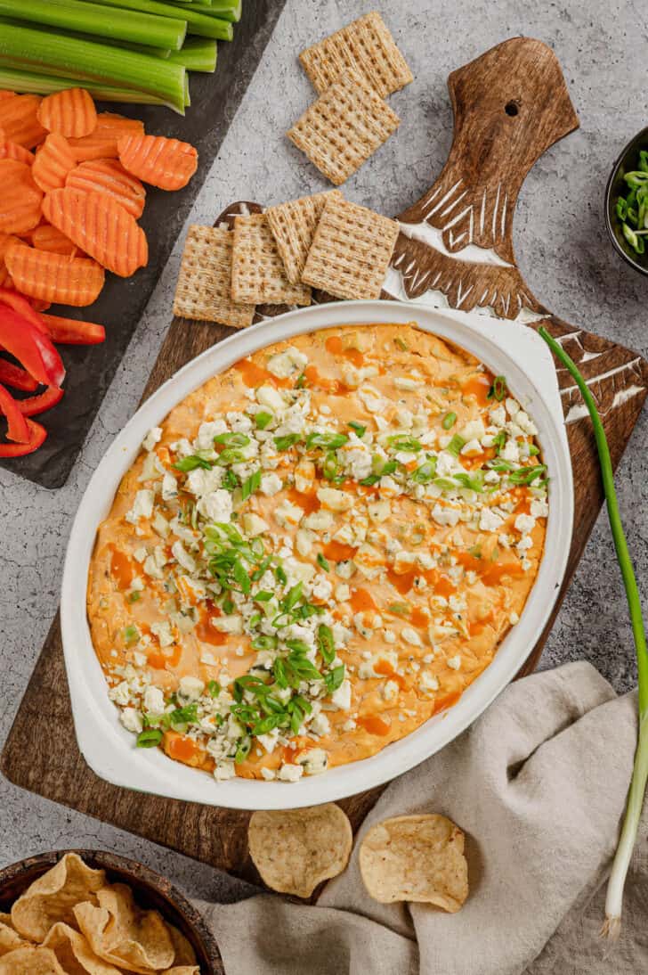 A baking dish full of healthy buffalo chicken dip on a cutting board, with crackers, cut veggies and tortilla chips alongside the dish.