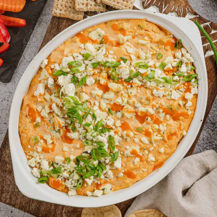 A baking dish full of healthy buffalo chicken dip on a cutting board, with crackers, cut veggies and tortilla chips alongside the dish.