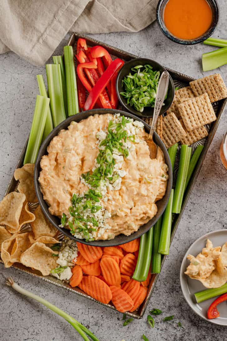 A bowl of healthy buffalo chicken dip on a platter with cut veggies, crackers and tortilla chips.