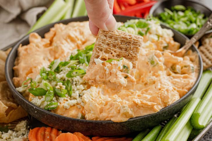 A large bowl of buffalo chicken dip made with Greek yogurt, with a hand dipping a wheat cracker into it.