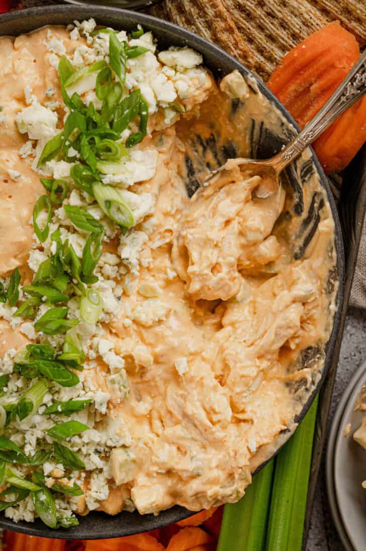 A spoon digging into a bowl of a cheesy orange mixture. The dip is garnished with blue cheese and green onions.