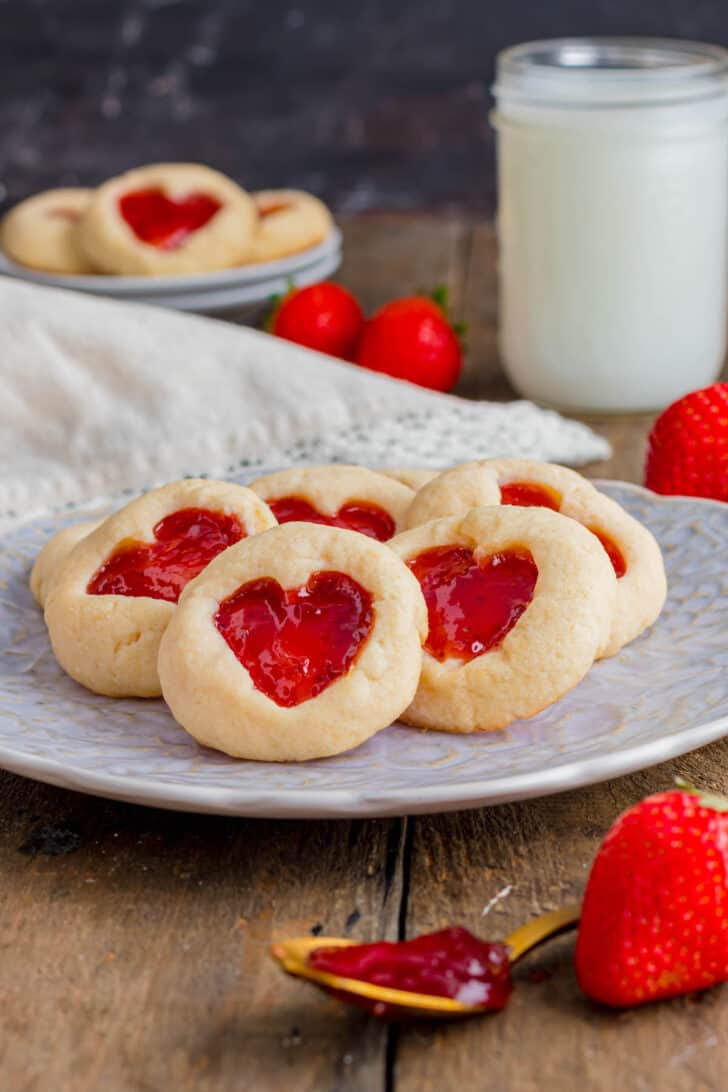 Shortbread heart jam cookies on a decorative plate with a glass of milk and fresh strawberries in the background.