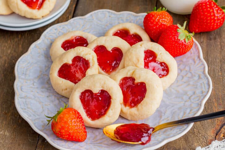 Heart thumbprint cookies filled with strawberry jam on a lacy plate.