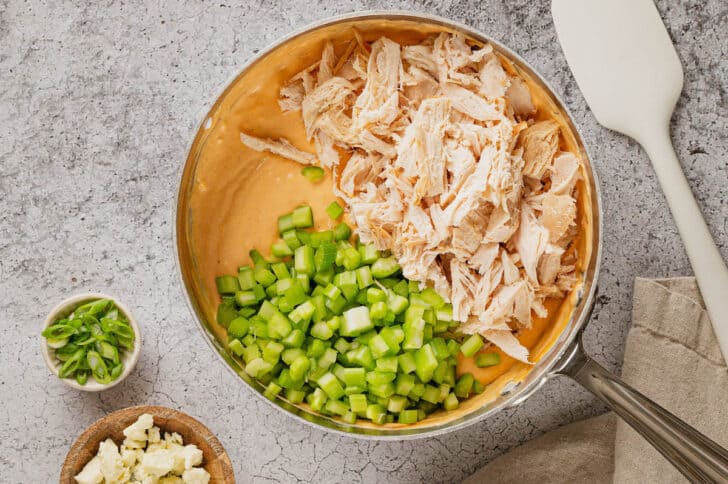 A saucepan filled with a creamy buffalo sauce mixture, topped with shredded chicken and chopped celery.