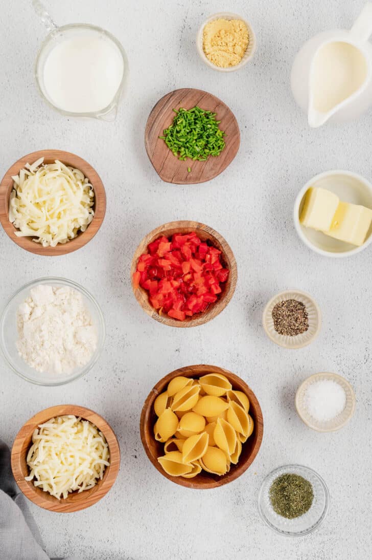 Ingredients laid out on a light surface, including milk, butter, shredded cheese, diced red peppers, pasta and seasonings.