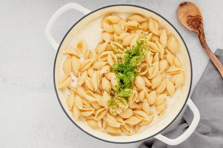 A white Dutch oven filled with a cream sauce topped with cooked pasta shells and chopped chives.
