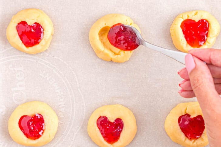 A woman's hand filling heart thumbprint cookies with red jam.