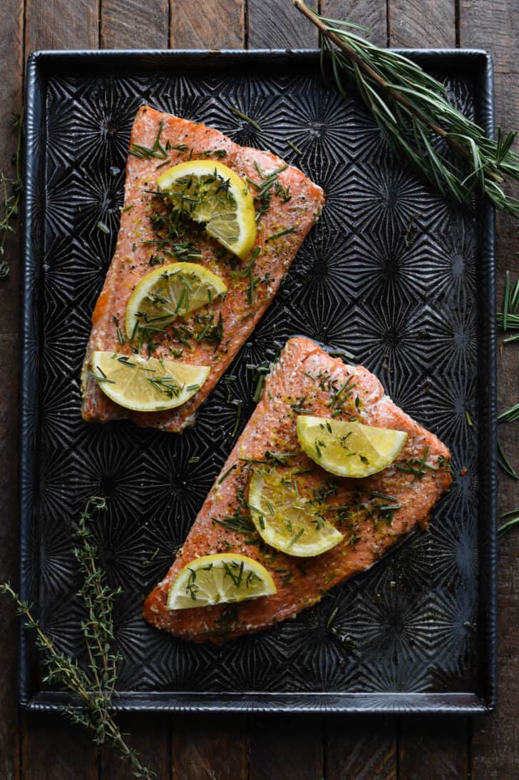 Baked sockeye salmon covered in herbs and lemon slices on a textured baking pan.