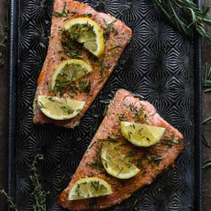 Baked sockeye salmon covered in herbs and lemon slices on a textured baking pan.