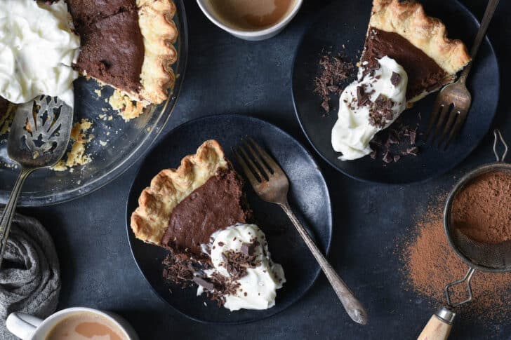 Two slices of brownie pie on small dark plates, topped with whipped cream and chocolate shavings.