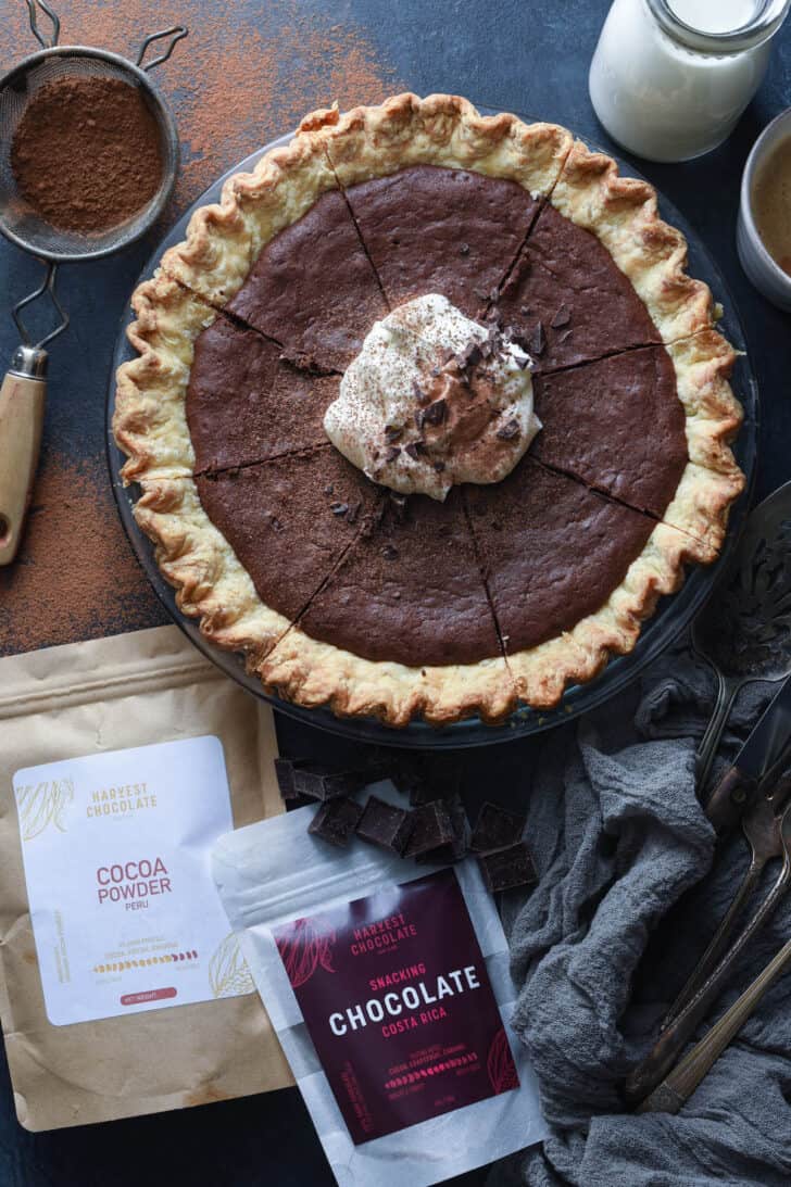 A chocolate tart topped with whipped cream alongside bags of Harvest Chocolate snacking chocolate and cocoa powder.