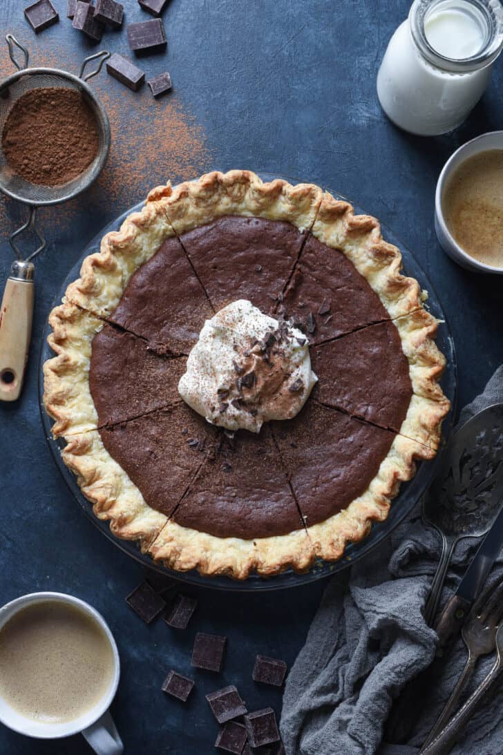 A brownie pie topped with whipped cream and chocolate shavings, on a table with mugs of coffee and milk.