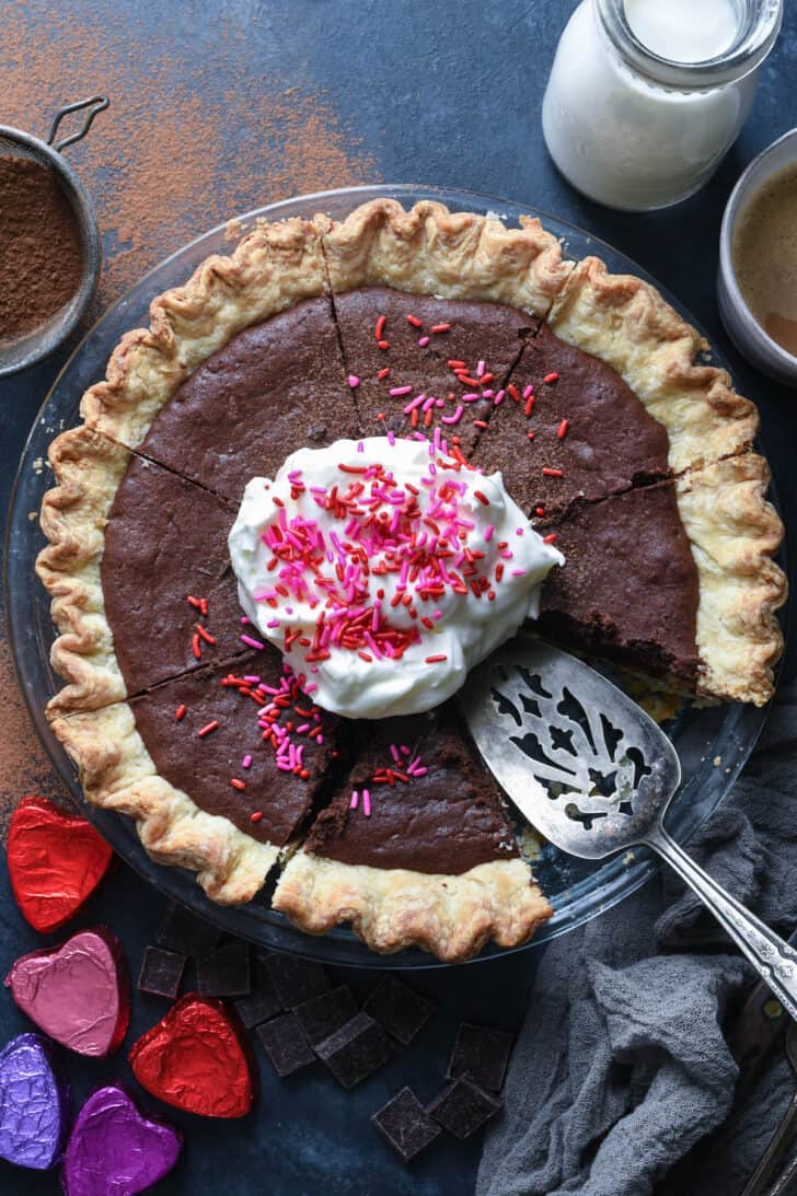 A chocolate brownie pie topped with whipped cream and pink and red spinkles, on a table with foil wrapped chocolte hearts.