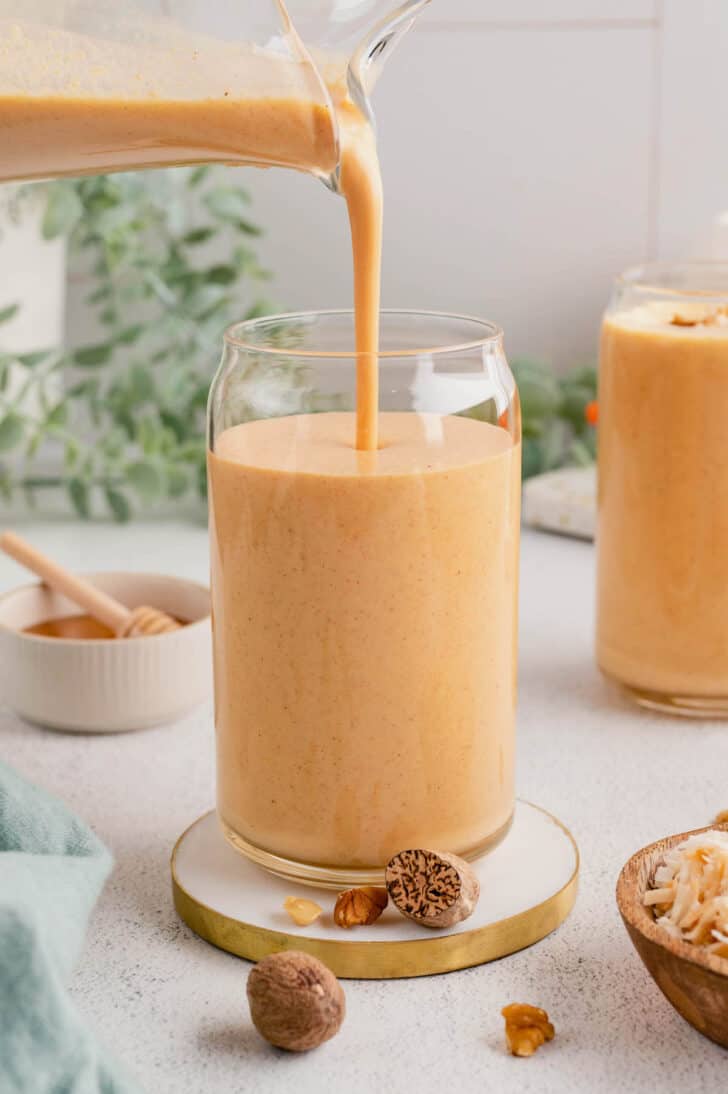 A carrot smoothie being poured out of a glass pitcher into a small glass.