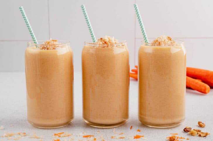 Three carrot cake smoothies lined up in a row, topped with coconut and garnished with light blue decorative paper straws.