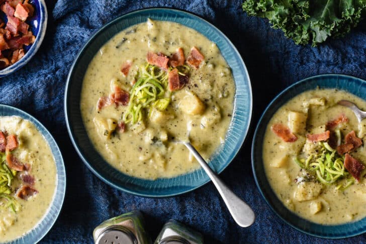 Three blue bowls on a blue linen, filled with cabbage and potato stew garnished with sliced leeks and crispy bacon.